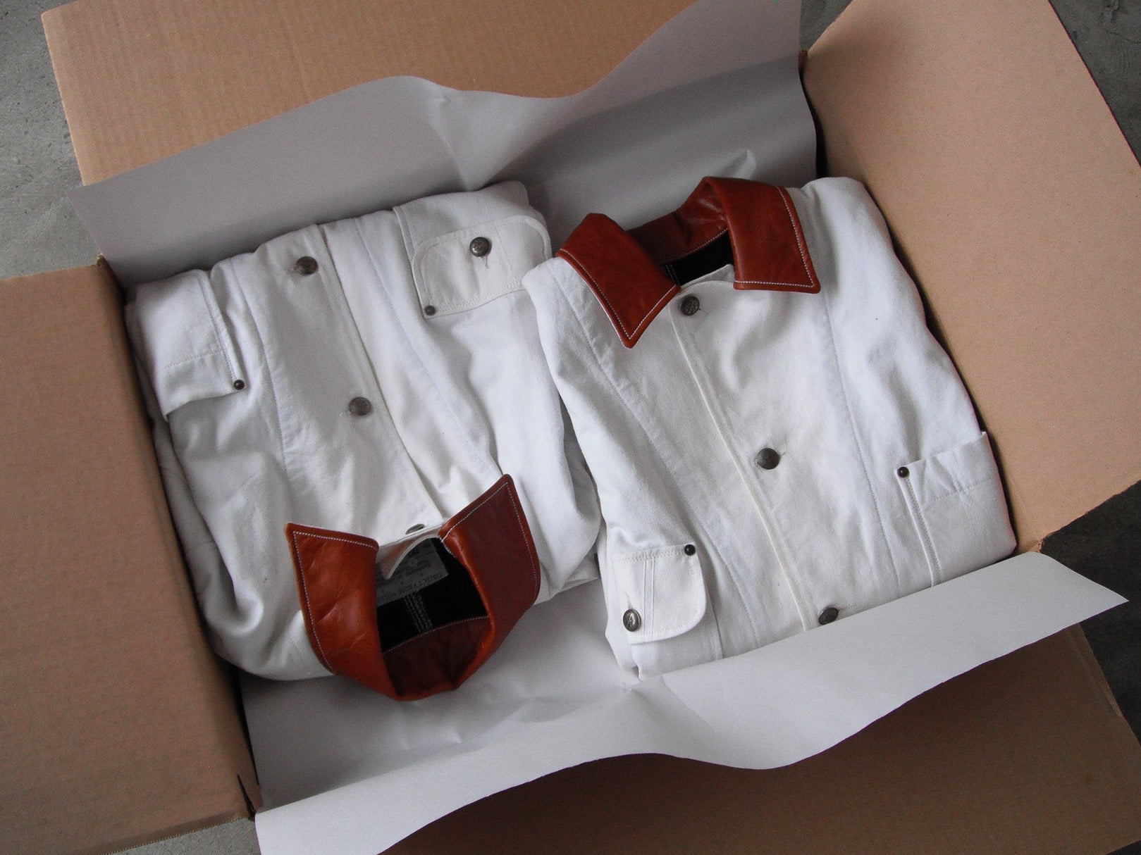 upcycled white jackets with brown leather collar in a cardboard box on a concrete floor.